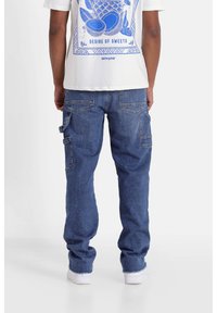 Pantalones cargo de mezclilla azul con múltiples bolsillos laterales, diseño de pierna recta y dobladillo deshilachado. Combinados con una camiseta gráfica blanca.