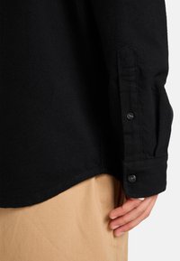 Zwarte button-up shirt van een gestructureerde stof, met een gebogen zoom en knoopmanchetten, gecombineerd met lichtgekleurde broek.