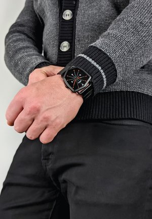 Sort sekskantet ur med orange time markører og sort rem, båret på en hånd. Personen er klædt i en grå og sort sweater.