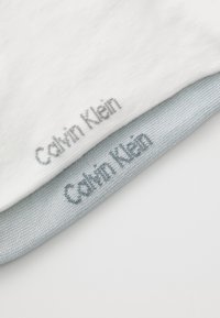 Két szövet címke található, rajtuk a "Calvin Klein" felirattal. A felső címke fehér, míg az alsó címke textúrázott világosszürke.