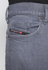 Vaqueros de mezclilla gris con un diseño clásico de cinco bolsillos, costuras visibles, remaches de metal y una etiqueta con "De Industry" en el bolsillo trasero.