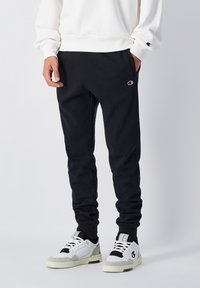 Pantalones de chándal negros con cintura elástica, dos bolsillos laterales y un pequeño logo. Complementados con una sudadera blanca y zapatillas de deporte en blanco y negro.