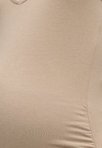 Κοντινό πλάνο ενός ατόμου που φοράει μπεζ, ελαστικό τοπ με ελαφρύ σούρωμα στο πλάι, υποδηλώνοντας εγκυμοσύνη ή περίγραμμα σώματος.