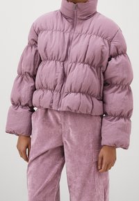 Puffig lavendelfärgad jacka med hög krage, dragkedja fram och vida ärmar; har en skrynklig struktur och elastiska muddar. Kombinerad med matchande velourbyxor.