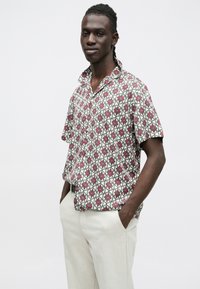 Koszula z krótkim rękawem ozdobiona kwiatowym wzorem w czerwieni i zieleni na beżowym tle, o swobodnym kroju i kołnierzyku.