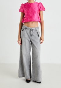 Haut rose court avec un motif floral texturé, associé à un jean gris à jambes larges avec ceinture et poches. Chaussures noires à bouts pointus.