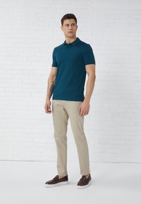 Teal polo skjorte med korte ermer og tre-knappers lukking, kombinert med beige bukser og brune slip-on sko, stående mot en hvit vegg.