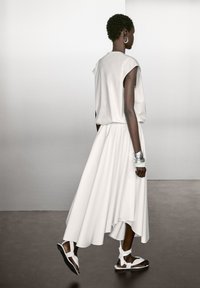 White sleeveless top with draped design, high-low hemline, and flowing skirt. Paired with silver bracelets and white sandals. Smooth texture.