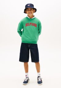 Αγόρι που χαμογελά με τα χέρια στην πλάτη, φορώντας πράσινο φούτερ Hilfiger, μπλε καπέλο τύπου bucket, σκούρα τζιν σορτς, λευκές κάλτσες και μπλε αθλητικά παπούτσια.