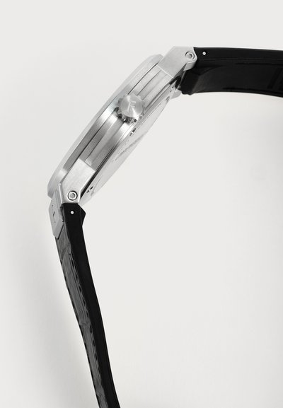 Vue de côté d'une montre-bracelet argentée avec un bracelet en cuir noir texturé et une couronne cannelée marquée de la lettre F.