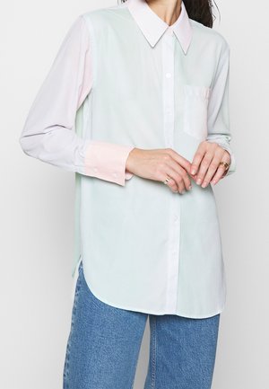 Vrouw draagt een losse pastelgekleurde overhemd met contrasterende manchetten en kraag, gecombineerd met blauwe jeans, handen samengevouwen ter hoogte van de taille.