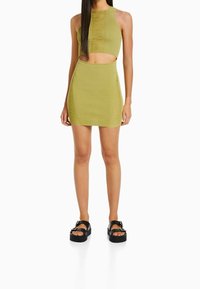 Světle zelený dvoudílný set obsahující přiléhavý crop top s texturovaným předním dílem a krátkou sukni s bočními výřezy, doplněný černými sandály na platformě.