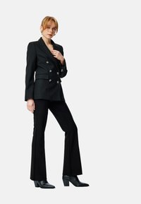 Blazer noir à double boutonnage avec des boutons argentés, associé à un pantalon noir ajusté et des bottes à talons pointues, se tenant contre un fond blanc.