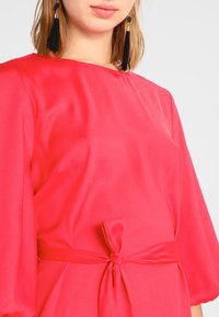 Světle růžové šaty s hladkou strukturou, mají vázání v pase a krátké nabírané rukávy. Jsou nošeny černé třásňové náušnice.