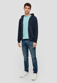 Sudadera con cremallera en color marino y cordones, sobre una camiseta de color azul claro, complementada con jeans azules y zapatillas blancas. Estilo casual y ajuste cómodo.