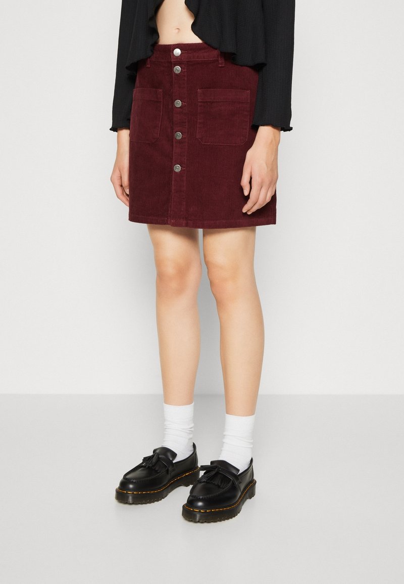 Femme portant une jupe boutonnée en velours côtelé bordeaux avec de grandes poches avant, des mocassins noirs, des chaussettes blanches et un haut noir court.