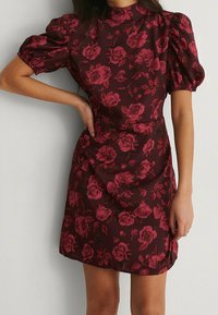 En person som bär en vinröd blommig klänning med puffärmar poserar mot en ljus bakgrund med händerna på höfterna.