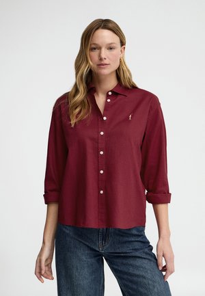 Femme aux longs cheveux blonds porte une chemise bordeaux boutonnée aux manches retroussées et un jean bleu, debout devant un fond blanc uni.