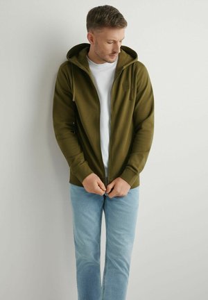 Mężczyzna w oliwkowozielonej bluzie rozpinanej na zamek i białej koszulce, zapinający zamek, stojący na tle jednolitego jasnoszarego tła.