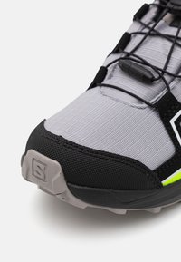 Gray and black athletic shoe with textured fabric, reinforced toe, and thin black laces. Features a bright green accent and rugged sole.