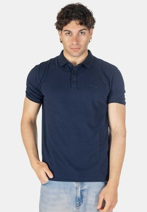 Giovane uomo con capelli ricci e scuri, che indossa una polo blu navy e jeans azzurri, in piedi davanti a uno sfondo chiaro semplice.