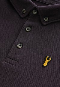 Donkerpaarse polo met textuurstof, drie knopen en een gele geborduurde kreeftenlogo op de linkerborst.