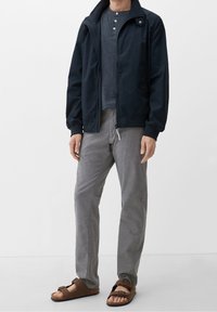 Marinblå zip-up jacka, grå henley-tröja, rutiga grå byxor och bruna sandaler. Jackan har ribbade muddar och en ställbar krage.