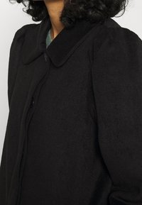 Abrigo negro de mezcla de lana con cierre de botones, cuello redondeado y mangas abullonadas, que presenta una textura suave y líneas limpias.