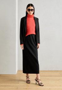 Black tailored blazer over a red ribbed turtleneck, paired with a black maxi skirt and strappy black heels. Smooth textures, modern design.
