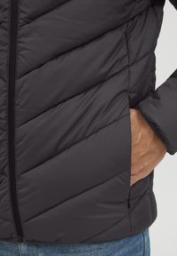 Jachetă puffer neagră cu un model țesut pe diagonală, închidere cu fermoar și buzunare laterale. Materialul pare neted și ușor.