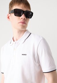 Occhiali da sole neri oversize con montatura angolare; abbinati a una polo bianca con un logo nero e un colletto a contrasto.
