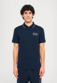Tamsiai mėlyna polo marškinėliai, pagaminti iš minkštos medžiagos, su trumpu užtrauktuku, kontrastingomis auksinėmis detalėmis ir EA7 logotipu kairiajame krūtinės srityje.