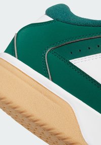 Grön och vit sportskor med en texturerad ovandel, gummisula och kontrasterande beige yttersula, med sömmar och perforeringar för andningsförmåga.