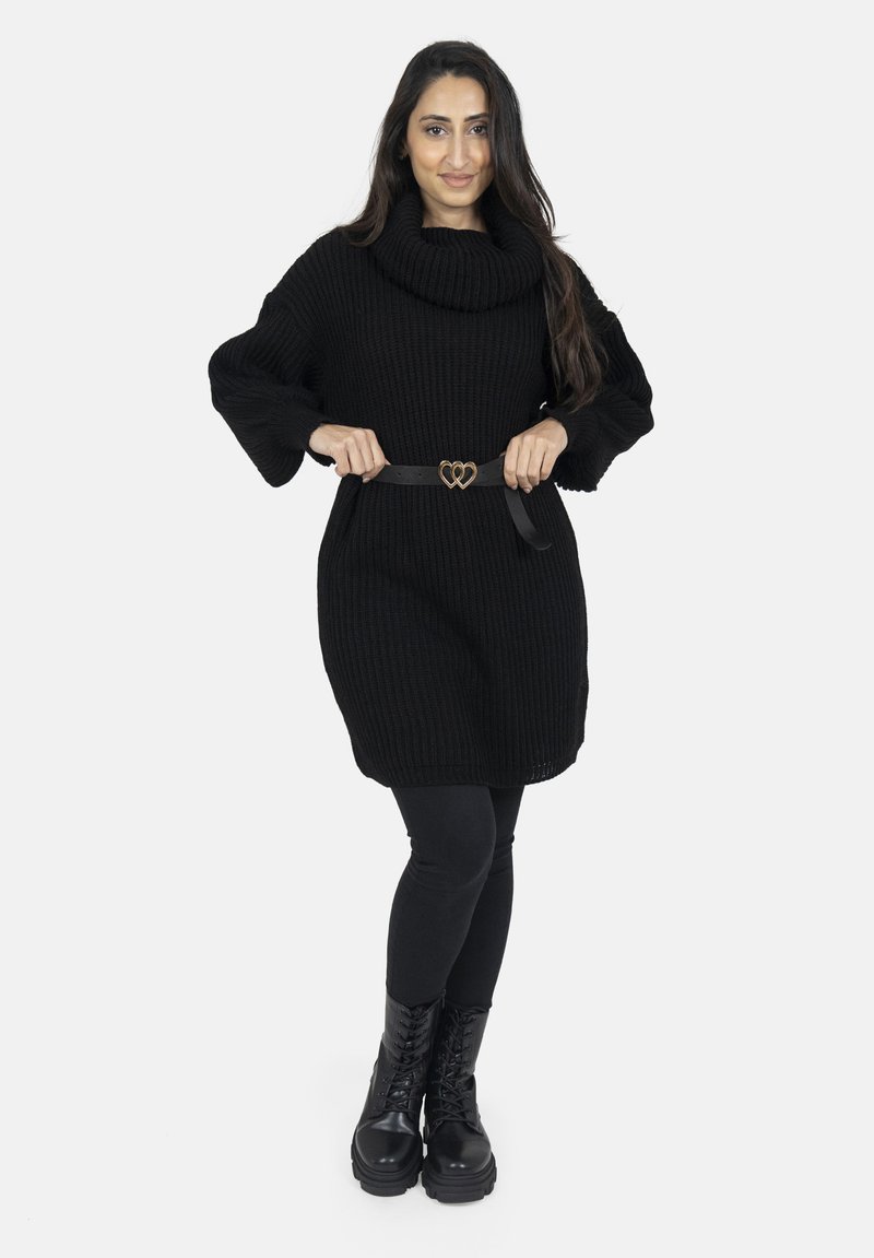 Pull en tricot noir surdimensionné avec un col cheminée, accompagné d'une ceinture noire avec une boucle en forme de cœur, de leggings noirs et de bottes de combat à lacets.