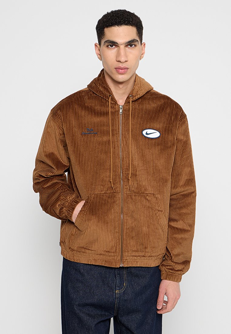 Hombre con una sudadera con cremallera de pana marrón y el logo de Nike, con las manos en los bolsillos, de pie frente a un fondo claro y liso.