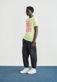 Φωσφοριζέ κίτρινη μπλούζα με γραφικό σχέδιο, συνδυασμένη με μαύρο παντελόνι φόρμας και λευκά αθλητικά παπούτσια. Το μοντέλο φοράει μαύρα γυαλιά ηλίου· απλό, καθημερινό ντύσιμο.