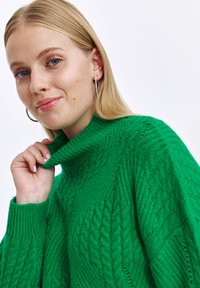 Zielony sweter z golfem wykonany na drutach, z teksturowanym wzorem w warkocze. Materiał wydaje się gruby i przytulny, z prążkowanymi mankietami i kołnierzem.