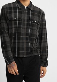 Camicia a zip a quadretti nera con linee grigie, due tasche sul petto con bottone e colletto. Tessuto strutturato, vestibilità rilassata e orlo elastico.