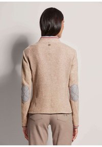 Beige strukturierte Jacke mit grauen Ellbogenflicken und roter Naht, taillierte Passform und Stehkragen mit roten Akzenten.
