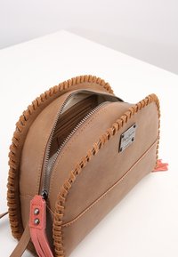 Kožená crossbody kabelka v tan barvě s kulatým tvarem, zdobená třásněmi, stříbrnými detaily a růžovým třásňovým akcentem. Se zipovým uzávěrem a vnitřní kapsou.