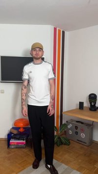 Bela športna majica s sivimi poudarki, črne hlače, rjavi čevlji. Tetovaže vidne na rokah. Preprost, moder dizajn v notranjem okolju.