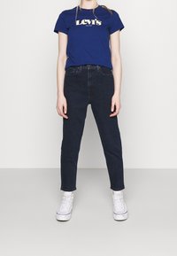 Person som har på sig en blå Levi's t-shirt, högmidjade svarta jeans och vita högstövelsneakers, står mot en ljus, enkel bakgrund.