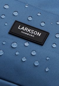 Modrá látková taška s kapkami vody, obsahující černou etiketu s nápisem "LARKSON HAMBURG" v bílém písmu. Texturovaný povrch s hladkým finišem.