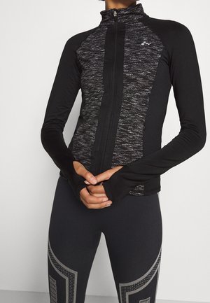 Vrouw draagt een zwarte sportjas met een volledige ritssluiting en een gestructureerd voorpaneel, gecombineerd met zwarte legging met grijze geometrische patronen. Handen zijn gevouwen voor haar.