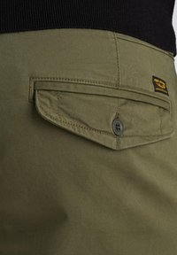 Pantalons cargo vert olive avec une poche arrière à rabat, fermeture à bouton et une étiquette noire avec une marque jaune. Tissu lisse et durable.