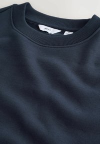 Prim-plan al unei hanorace de culoare albastru închis, având un decolteu cu nervuri și o etichetă cu textul "NEXT" și mărimea "M" vizibilă.