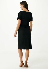 Vestito nero a maniche corte con una vestibilità rilassata, lunghezza al ginocchio e tessuto morbido. Abbinato a sandali con tacco marroni, che mostrano un design pulito e minimalista.