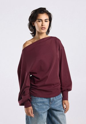 Young person with short dark hair wearing an off-shoulder burgundy sweatshirt and loose blue jeans, standing against a plain background.