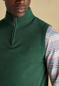 Vyras vilkintis žalią zip-up be rankovių megztinį ant marguoto langelio rašto ilgomis rankovėmis marškiniai, šviesiai smėlio spalvos fonas.