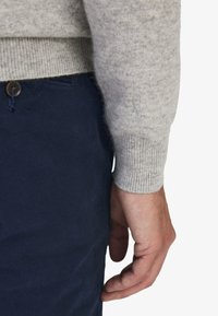 Grå ulltröja med ribbade manschetter, i kombination med marinblå byxor med en knappdetalj. Närbild som visar sidan av handen som vilar på byxorna.